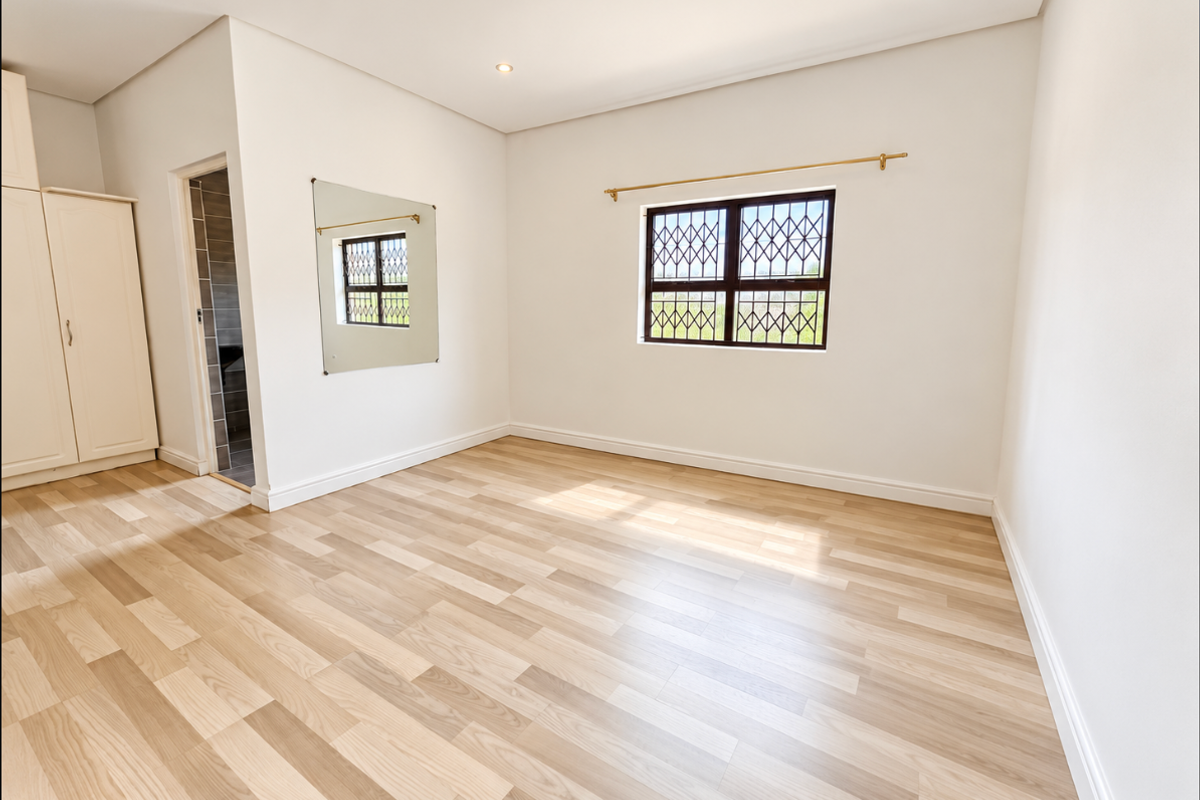A bright, empty bedroom featuring light wood-look laminate flooring, built-in wardrobe storage, and a window fitted with a security gate.