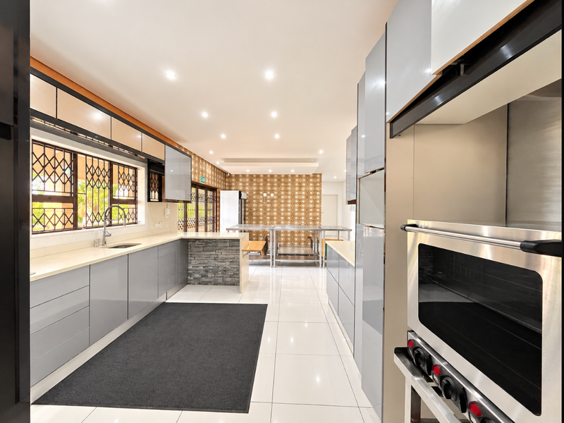 A spacious, modern kitchen featuring sleek grey cabinetry, stone countertops, integrated stainless steel oven, and bright recessed lighting with tiled flooring throughout.
