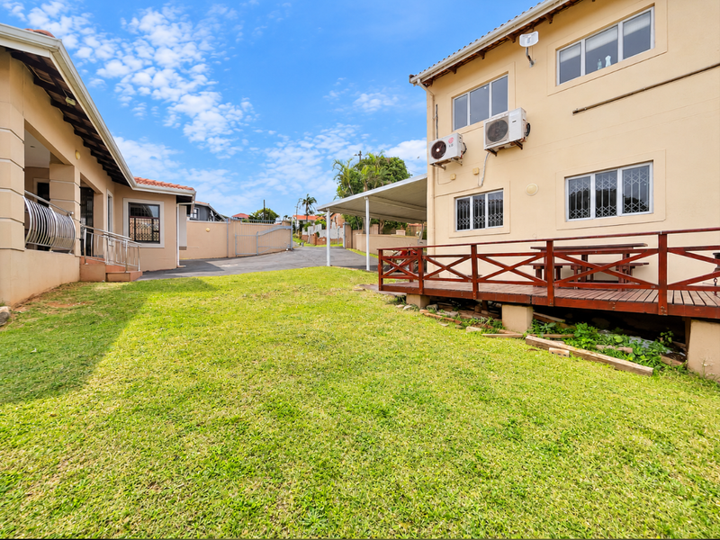 Rear exterior view featuring a lawn, a raised timber deck, and a paved driveway area with a carport.