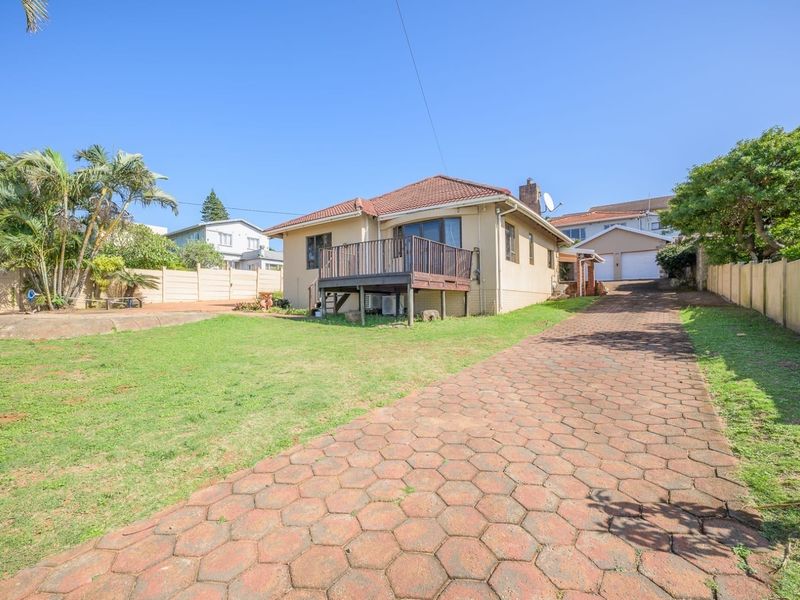 Rear view of the property featuring a raised timber deck, a large paved driveway, and a spacious garden area with boundary walling.