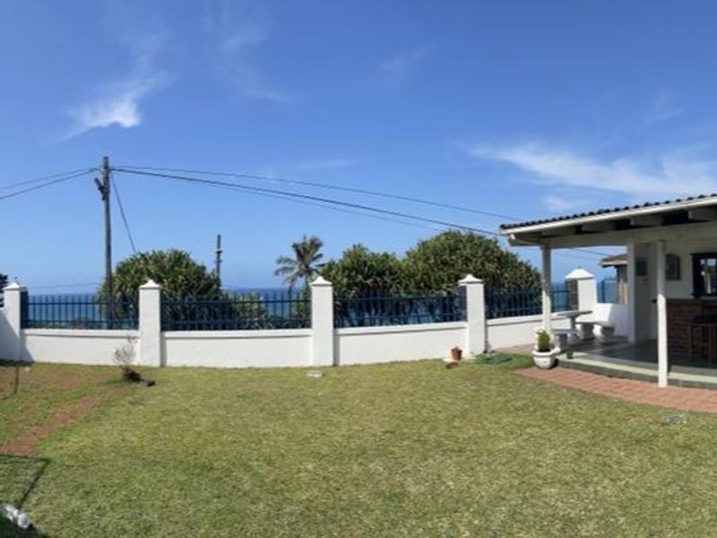 A rear garden area featuring a well-maintained lawn, a boundary wall with metal fencing, and a covered patio space with ocean views.