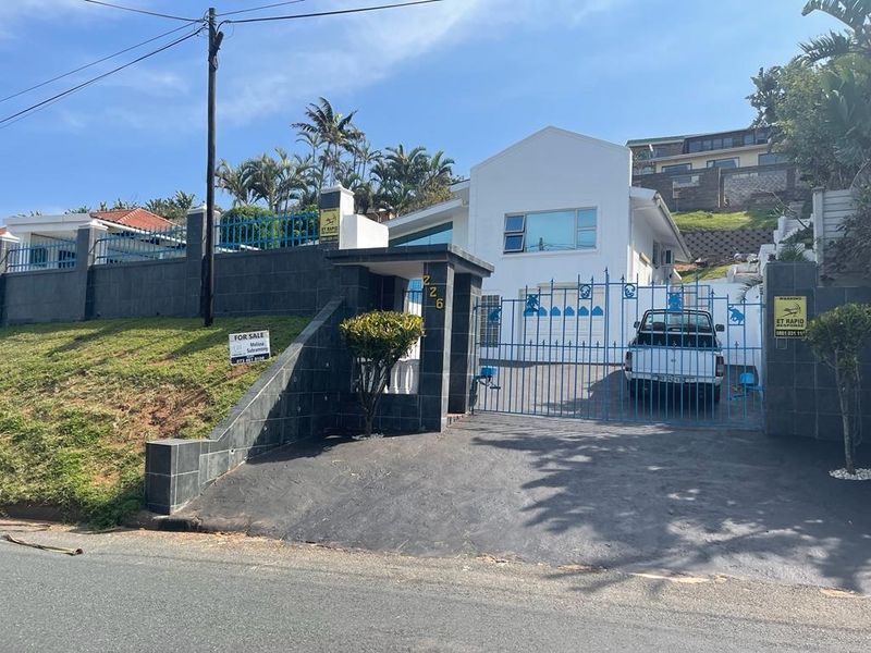 Front elevation of the property featuring a paved driveway, secure metal gate, tiled boundary walls, and a single garage entrance.