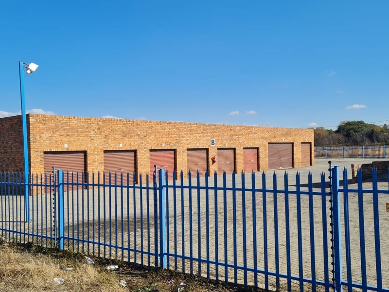 Industrial storage facility featuring a row of individual roller shutter units, secure perimeter fencing with electric security, and a paved driveway area.