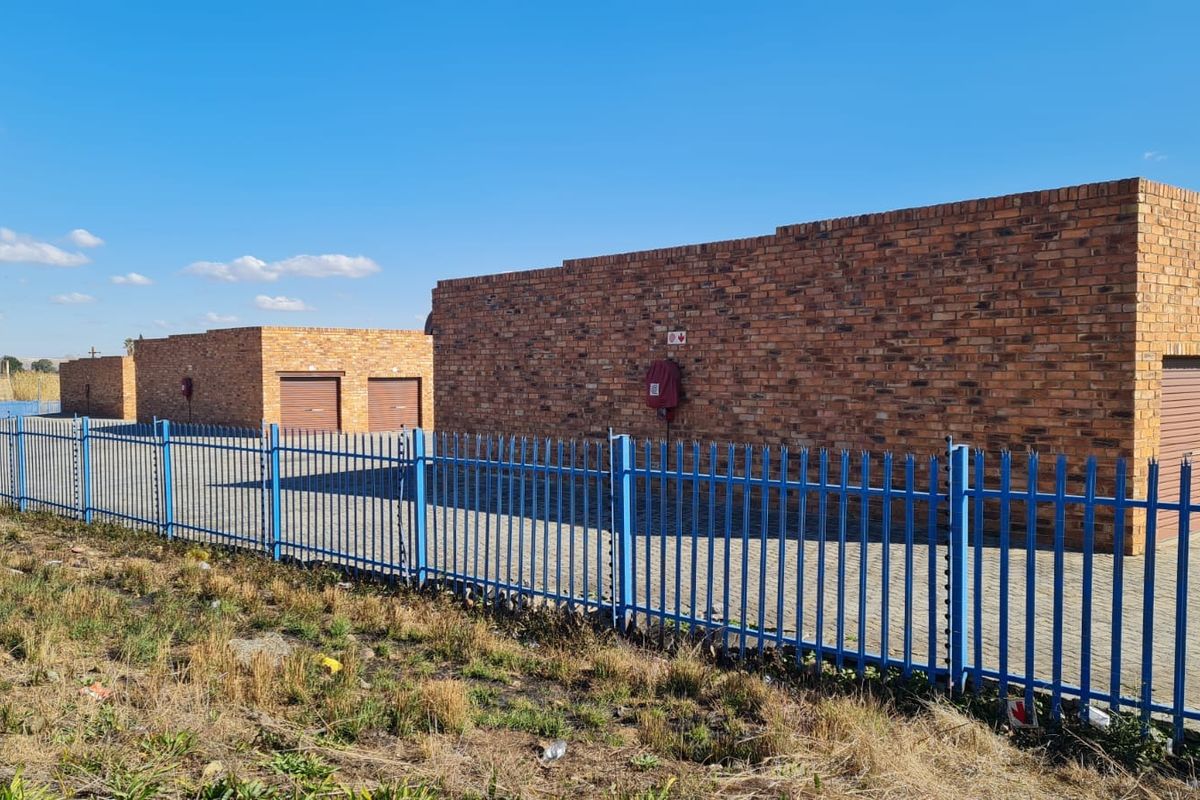 Industrial property featuring face brick construction, secure steel palisade fencing, paved driveway access, and multiple roller shutter doors for vehicle or storage entry.
