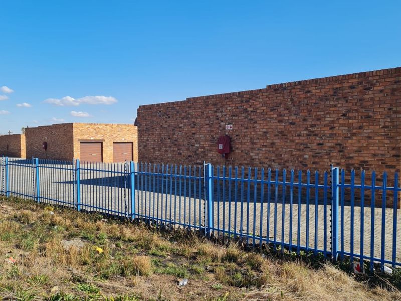 Industrial property featuring face brick construction, secure steel palisade fencing, paved driveway access, and multiple roller shutter doors for vehicle or storage entry.