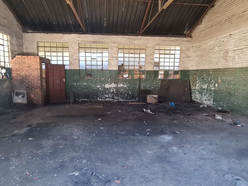 Spacious industrial utility area featuring exposed steel roof trusses, concrete flooring, and large steel-framed windows, requiring significant refurbishment.