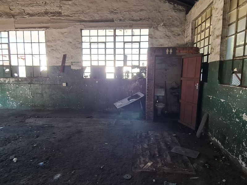 Spacious industrial utility area featuring exposed brickwork, steel-framed windows, concrete flooring, and an integrated toilet cubicle, requiring significant renovation and refurbishment.