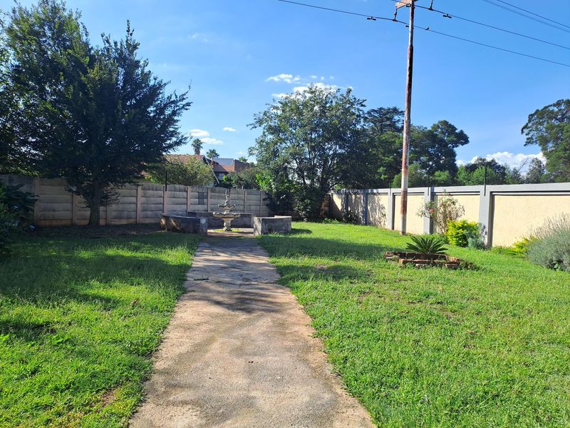 A spacious backyard garden featuring a central stone fountain, concrete pathway, and perimeter boundary walls.