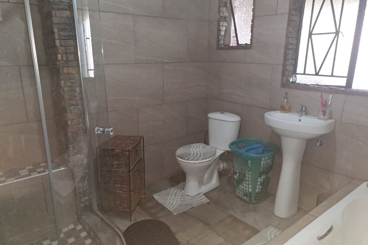 A functional bathroom featuring a glass-enclosed shower, pedestal basin, and toilet, finished with neutral wall tiles and a window with security bars.
