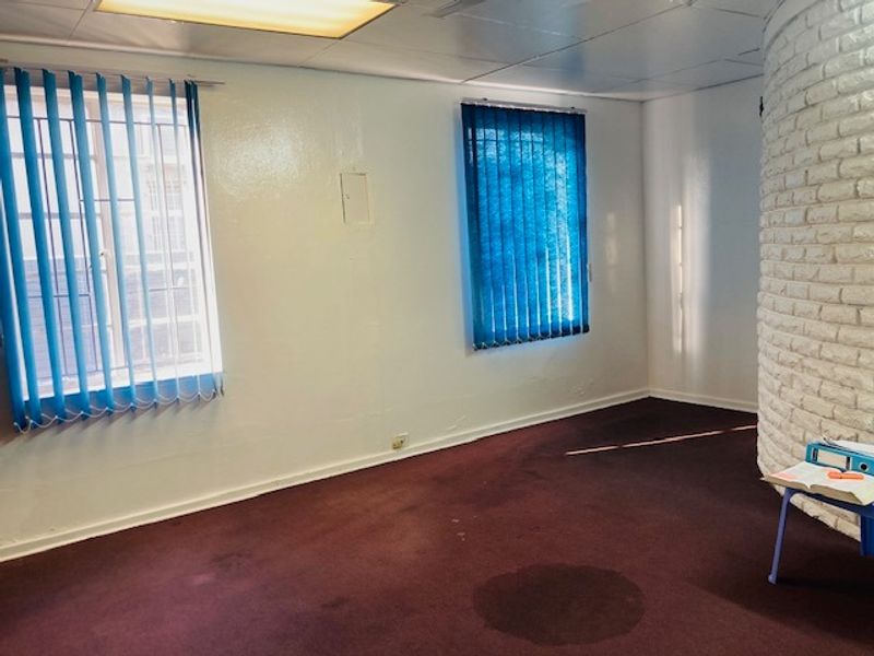 An empty living area featuring a curved white brick feature wall, large windows with vertical blinds, and carpeted flooring.