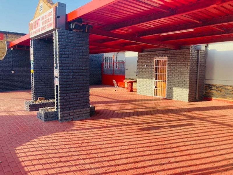 A paved front exterior area featuring a large covered carport, brick pillars, and secure gated access, suitable for parking or commercial use.