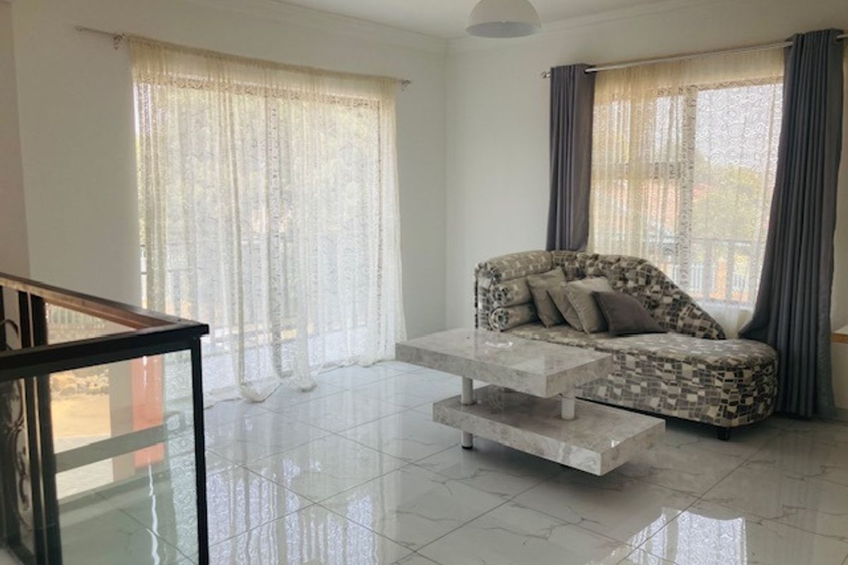 A bright living area featuring polished porcelain floor tiles, large windows with curtains, and a glass-panelled staircase railing.