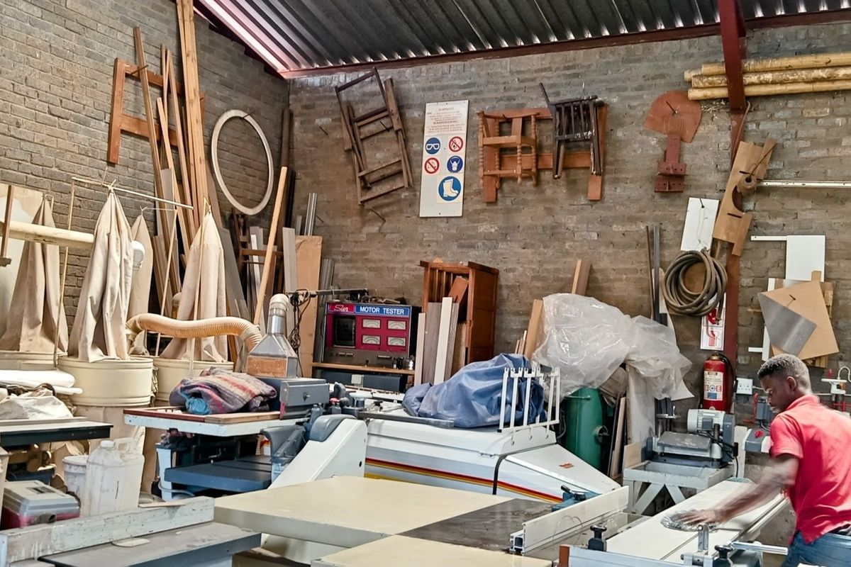 Industrial workshop space featuring exposed brick walls, high-pitched corrugated metal roofing, and steel structural trusses, currently utilized for woodworking operations.