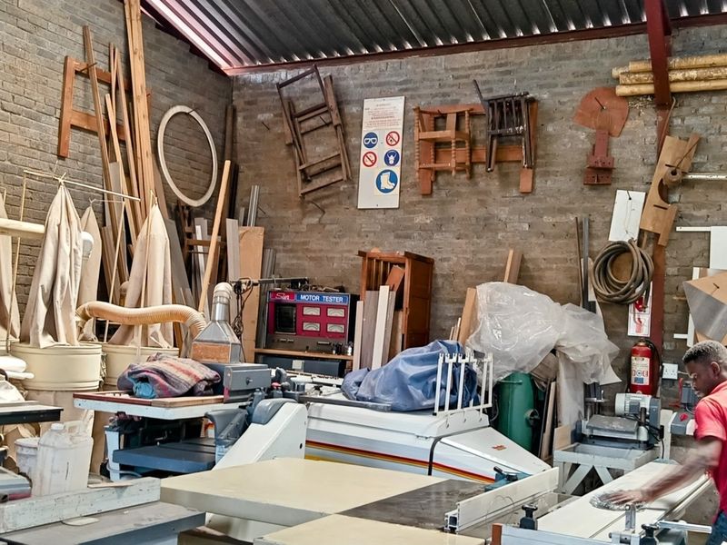 Industrial workshop space featuring exposed brick walls, high-pitched corrugated metal roofing, and steel structural trusses, currently utilized for woodworking operations.