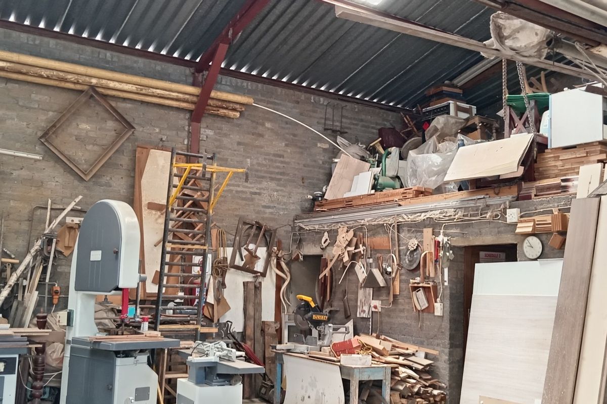 Industrial workshop space featuring exposed brick walls, corrugated metal roofing, and a mezzanine storage level, currently utilized for woodworking operations.