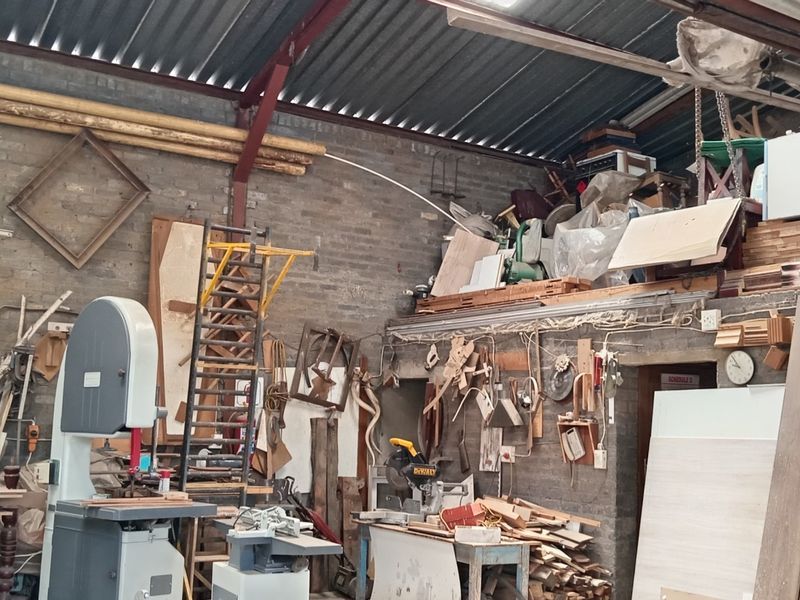 Industrial workshop space featuring exposed brick walls, corrugated metal roofing, and a mezzanine storage level, currently utilized for woodworking operations.
