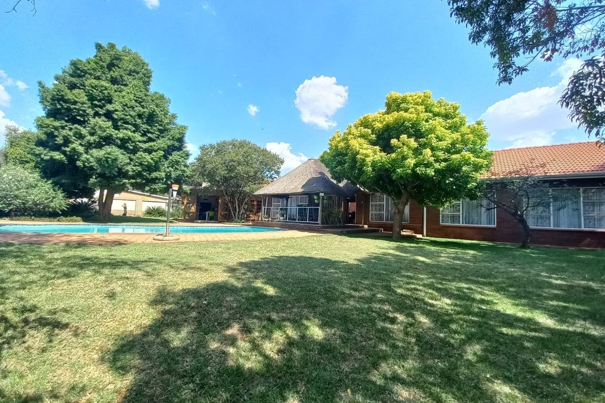 Spacious rear garden featuring a swimming pool, a traditional thatched lapa entertainment area, and established trees set against a brick-built home.