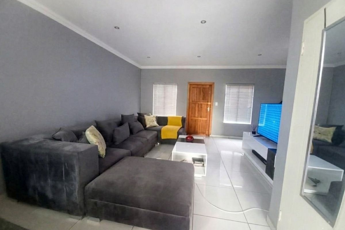 A spacious living room featuring neutral grey walls, polished tiled flooring, recessed downlights, and natural light through venetian blinds.