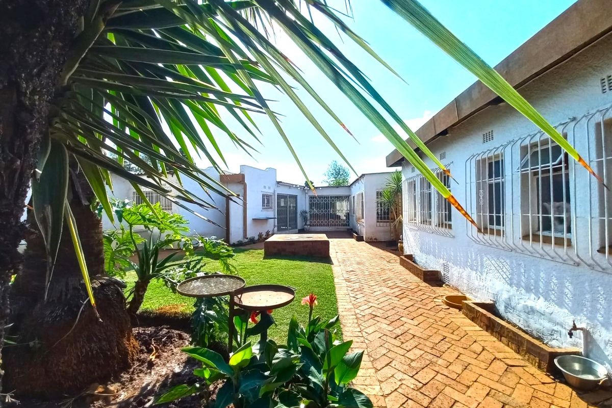 A rear exterior view featuring a brick paved walkway, a well-maintained lawn, and security bars on the windows of the main building structure.