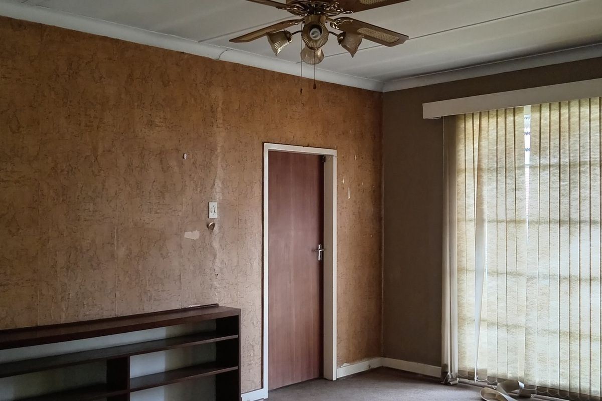 A vacant living room featuring a ceiling fan, built-in shelving, and vertical blinds, requiring cosmetic updates to the wall finishes.
