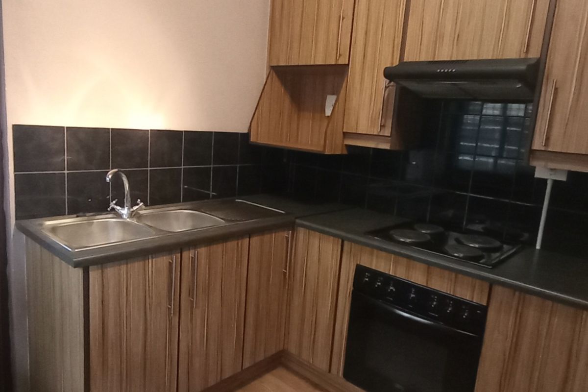 A functional kitchen featuring wood-look laminate cabinetry, a double bowl sink, black tiled splashback, and built-in electric cooking appliances.