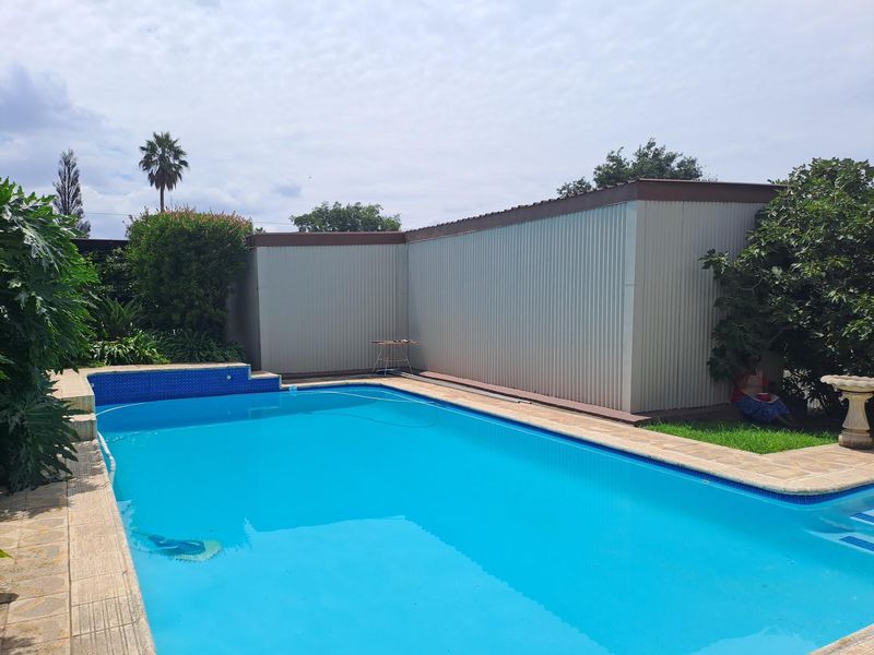 Outdoor swimming pool area featuring blue tiled coping, stone paving, and a large corrugated metal structure in the background.