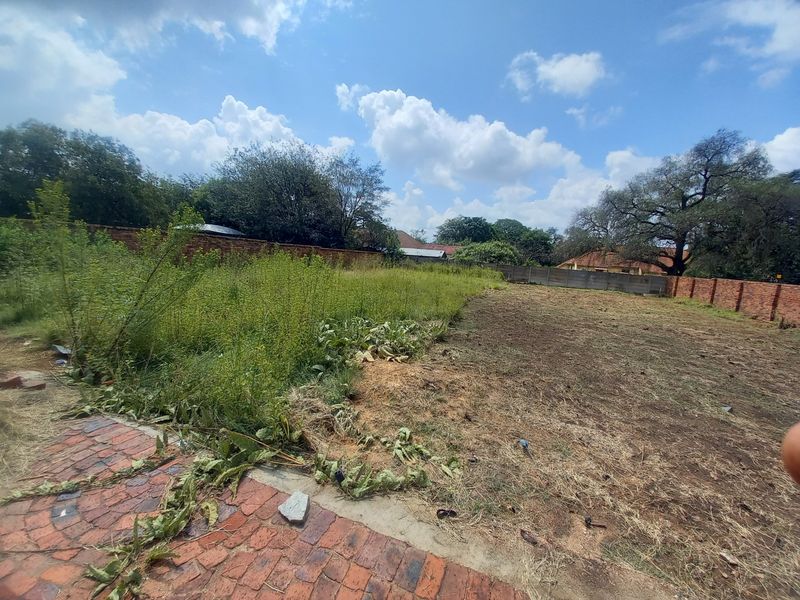 A vacant residential plot featuring a mix of cleared earth, overgrown vegetation, and perimeter boundary walls with existing brick paving in the foreground.