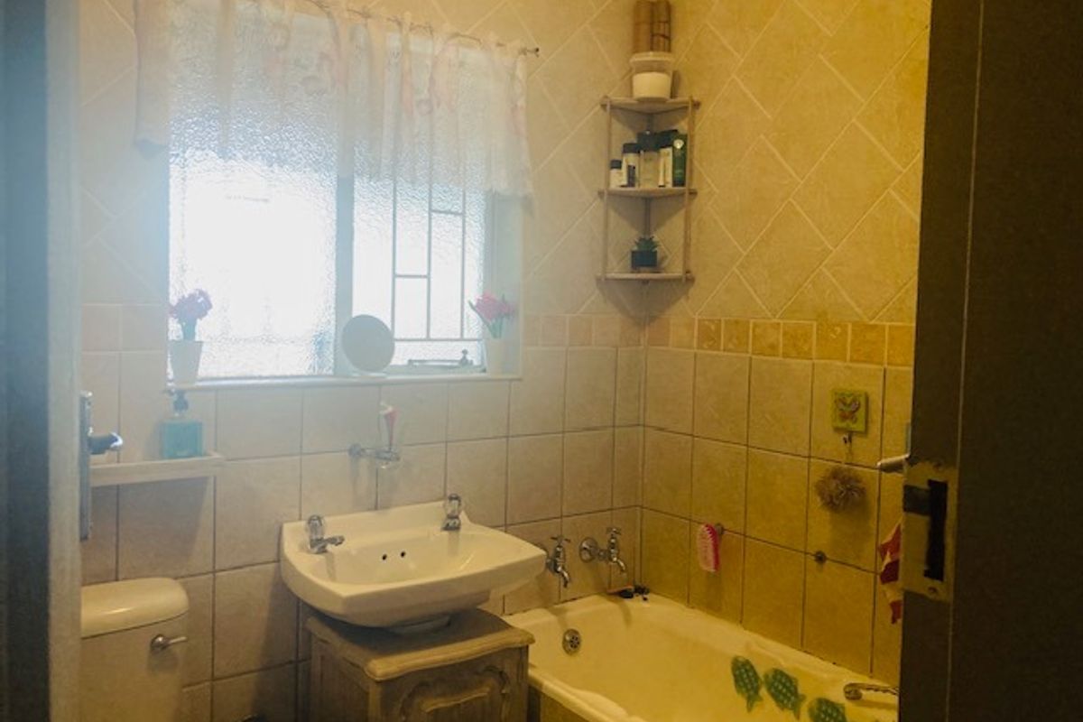A functional bathroom featuring a bathtub, pedestal washbasin, and toilet, finished with dated ceramic wall tiles and security bars on the window.