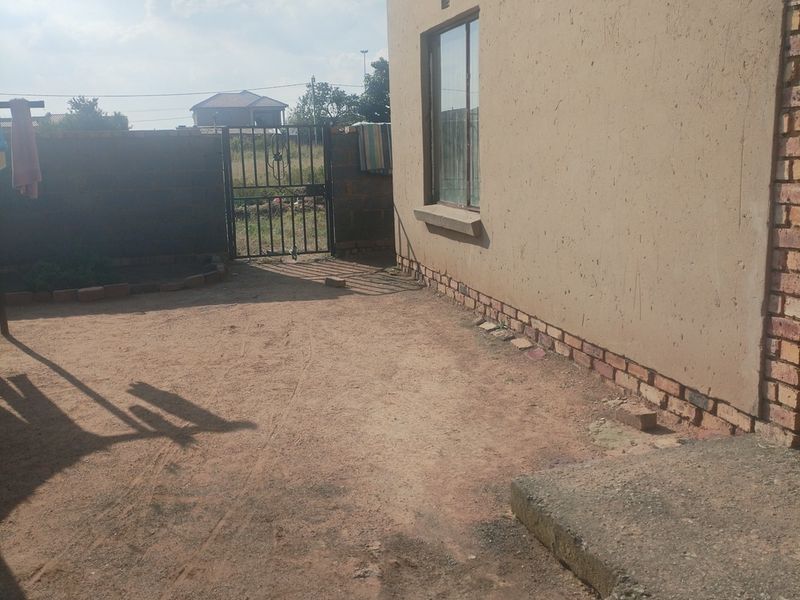 Rear exterior view of the property featuring a plastered wall, concrete step, and metal gate leading to an unpaved yard area.