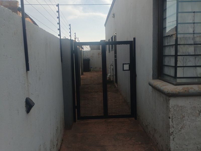 Side passage featuring a security gate, electric fencing, and a bay window with security bars, showing signs of exterior wall wear.