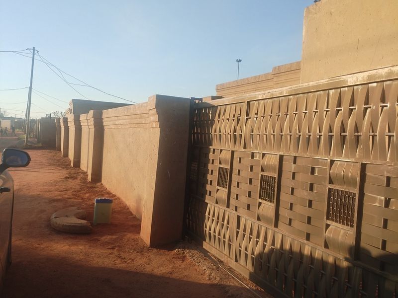 Front view of the property featuring a tall perimeter wall and a decorative metal sliding gate along an unpaved street.