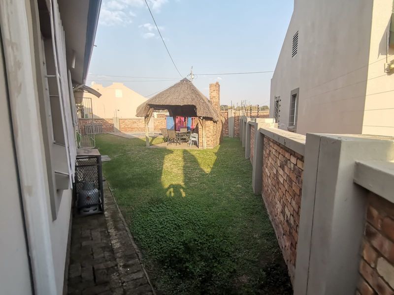 A rear garden area featuring a thatched lapa with a brick chimney, enclosed by brick boundary walls and a paved side walkway.