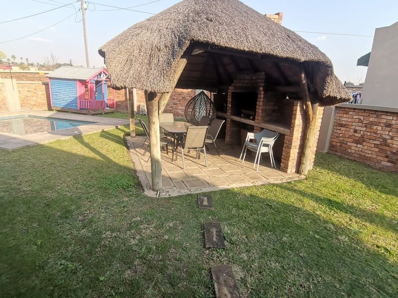 Outdoor entertainment area featuring a thatched lapa with a built-in brick braai, adjacent to a swimming pool and garden space.