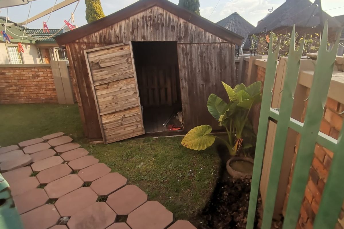A backyard area featuring a weathered wooden garden shed, paved walkway, and brick boundary wall with metal security fencing.
