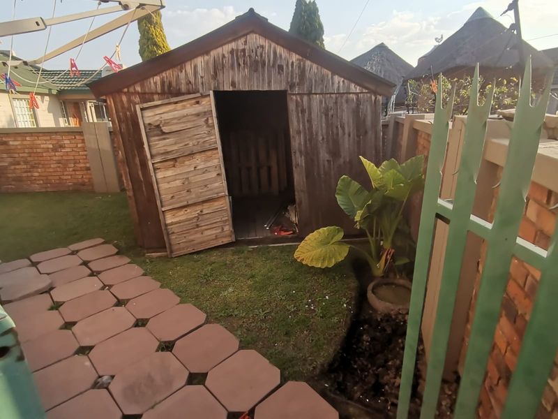 A backyard area featuring a weathered wooden garden shed, paved walkway, and brick boundary wall with metal security fencing.