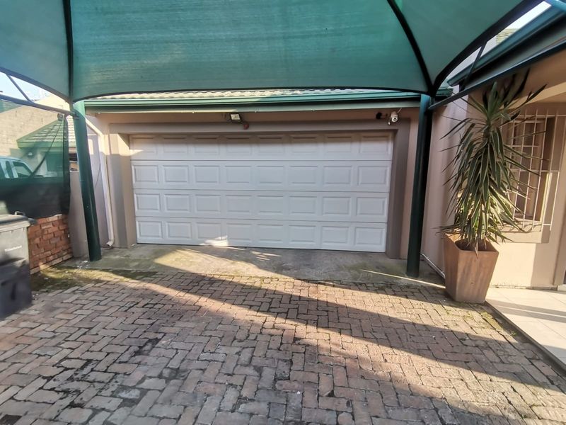 A single garage featuring a white sectional door, protected by a green shade cloth carport structure over a brick paved driveway.