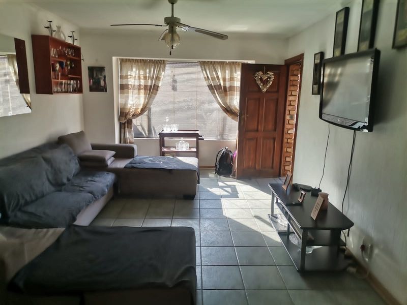 A living room featuring tiled flooring, a ceiling fan, and natural light through a window with blinds and a timber entrance door.