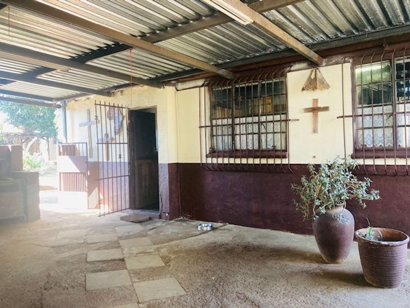 A covered outdoor patio area featuring a corrugated metal roof, security bars on windows, and paved flooring, providing a functional sheltered space.