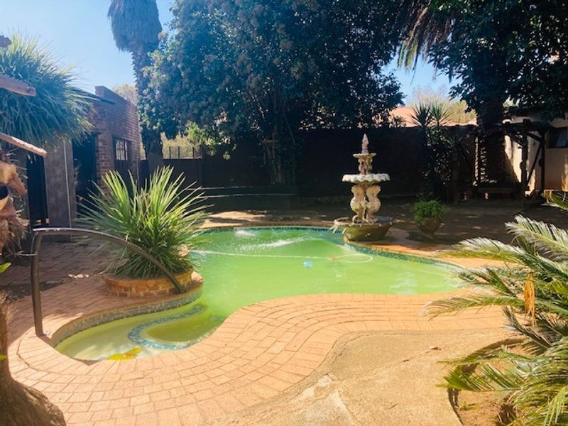 Outdoor pool area featuring a curved swimming pool with integrated water fountain and brick paved surround, set within a mature garden setting.