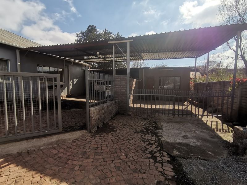 Front exterior view featuring a paved driveway, steel security gate, and a large corrugated metal carport structure.