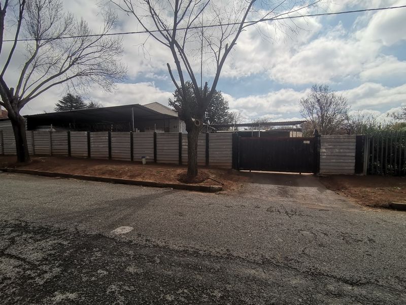 Street view of the property frontage featuring a precast concrete boundary wall, metal driveway gate, and an existing carport structure.