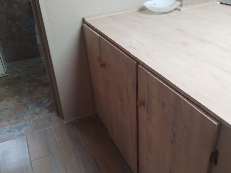 A clean kitchen area featuring light wood-look cabinetry, matching laminate countertops, and modern ceramic floor tiles, with an adjacent view into a tiled bathroom.