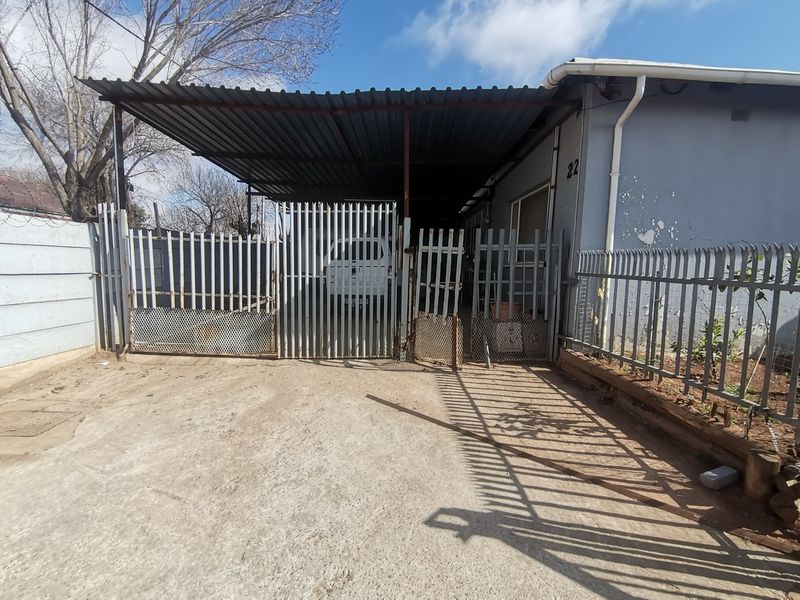 Front exterior view featuring a concrete driveway, metal carport, and steel palisade fencing with visible wear to the building facade.