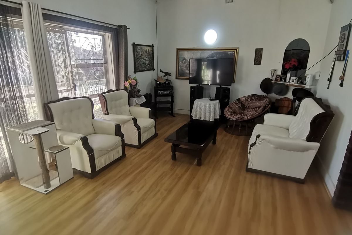 A living room featuring laminate flooring, ornate ceiling cornices, and a large window with security bars, offering a dated but functional living space.