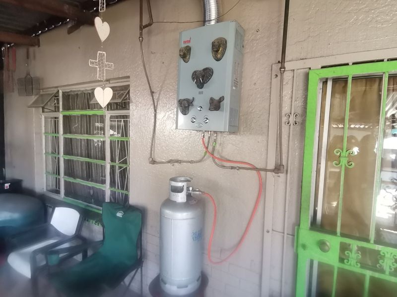 Outdoor utility area featuring a wall-mounted gas geyser with connected gas cylinder and security gate access.