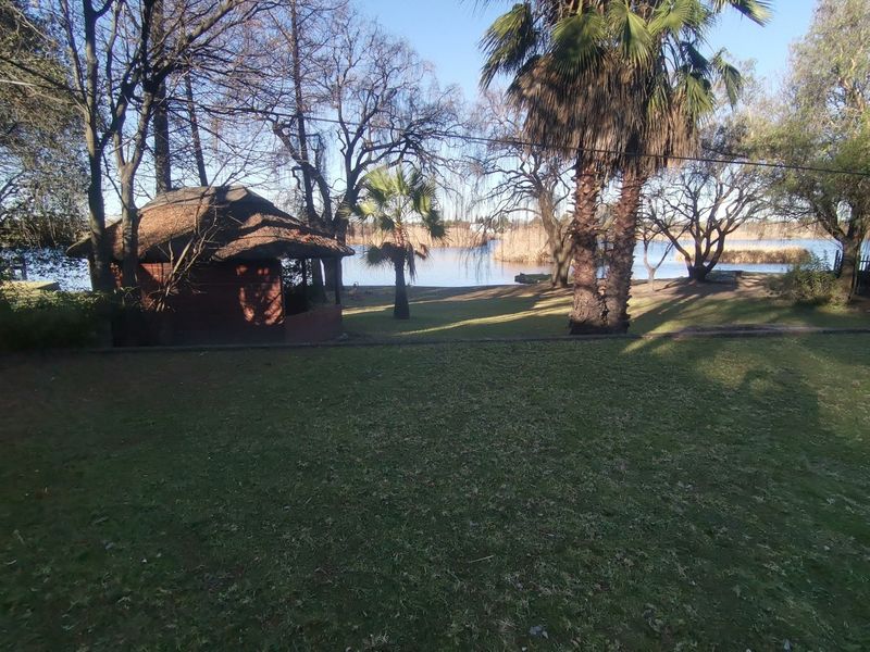 A spacious garden featuring a thatched gazebo, mature trees, and palm trees, with a scenic view of the nearby lake.