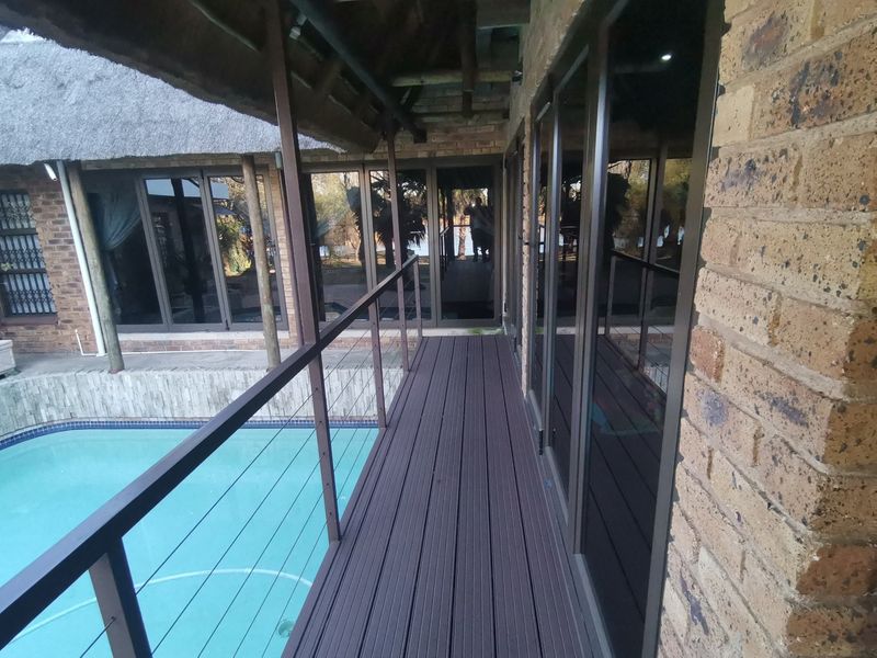 A raised composite deck walkway overlooking a swimming pool, featuring exposed brick walls, thatched roofing, and large sliding glass doors.