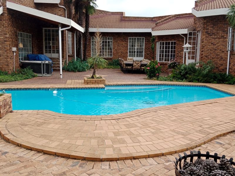 A spacious outdoor pool area featuring a swimming pool surrounded by brick paving, set against a face-brick house with a covered patio.
