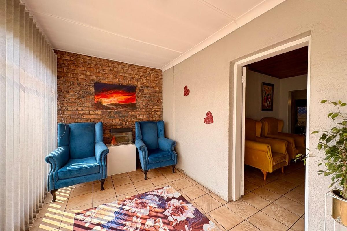A sunlit living area featuring an exposed brick feature wall, tiled flooring, and vertical blinds, providing a cozy space with direct access to an adjacent room.