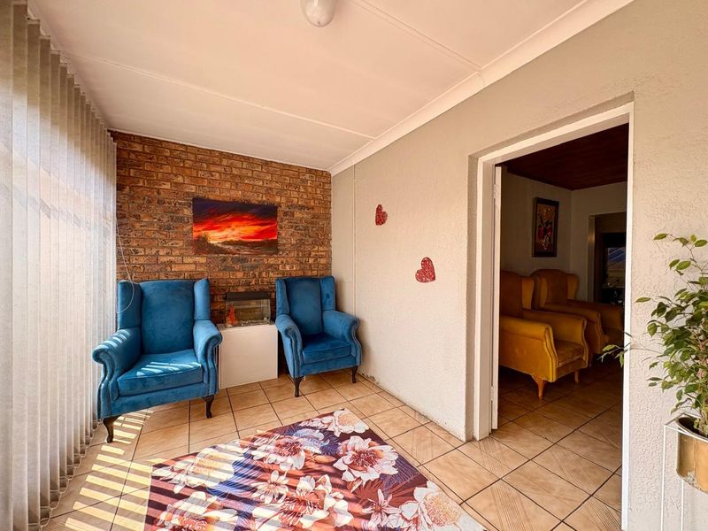 A sunlit living area featuring an exposed brick feature wall, tiled flooring, and vertical blinds, providing a cozy space with direct access to an adjacent room.